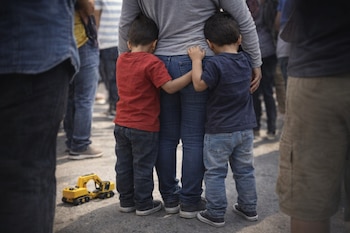 Dos niños, vistos de espaldas, se aferran a las piernas de una mujer. Un juguete excavadora amarillo yace en el suelo rodeado de gente.