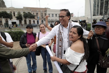 Un hombre con gafas y bufanda saluda a una mujer mientras un grupo de personas sonríe en una calle urbana con edificios y agentes de policía