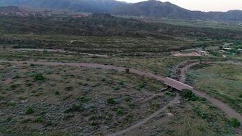 Vista aérea de un paisaje árido con vegetación dispersa, un camino de tierra, un puente de hormigón sobre un cauce seco y montañas al fondo