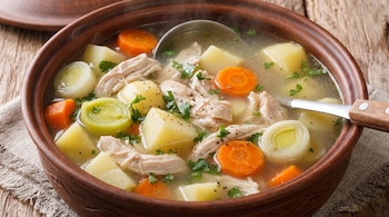 Primer plano de un tazón de cerámica marrón con caldo de pollo humeante, papas, zanahorias, puerros, pollo desmenuzado y perejil, con una cuchara. Superficie de madera.