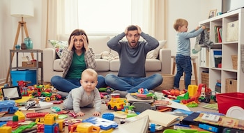 Padres con las manos en la cabeza, sentados en el suelo de una sala de estar desordenada. Un bebé gatea y un niño pequeño saca cosas de un estante.