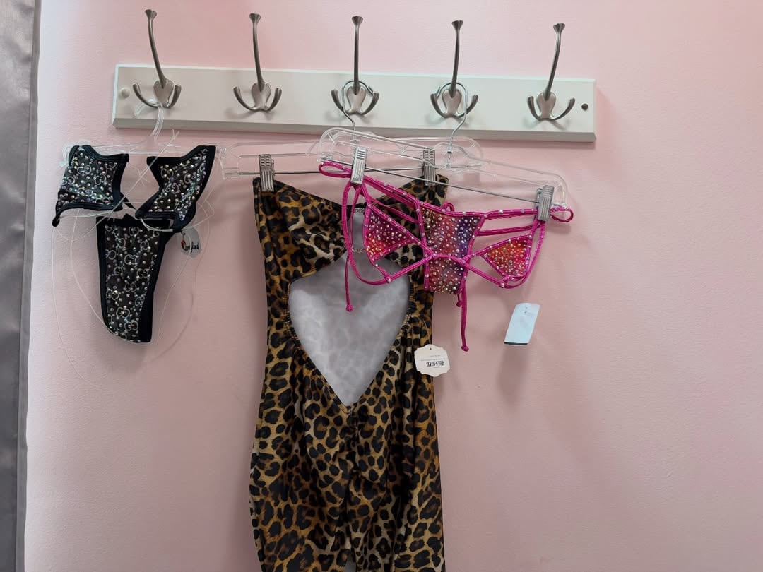 Variedad de prendas de moda playera, incluyendo un bañador negro con pedrería, un vestido animal print y un top de bikini rosa con brillantes, cuelgan en un perchero.