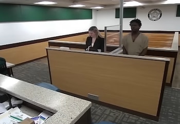 Hombre de tez oscura con uniforme beige y mujer rubia con blazer negro de pie en sala judicial con tabiques de madera y escudo en la pared