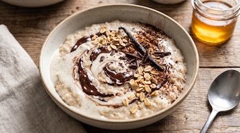 Un tazón con avena, chocolate derretido, cacao en polvo, copos de avena y una vaina de vainilla junto a una cuchara de metal.