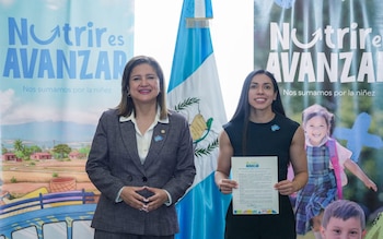 Dos mujeres de pie, una bandera de Guatemala, dos pancartas azules con el texto Nutrir es Avanzar, una de las mujeres sostiene un documento