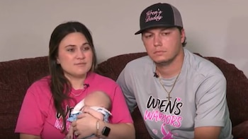Una mujer con camiseta rosa sostiene a un bebé junto a un hombre con camiseta gris y gorra negra en un sofá marrón