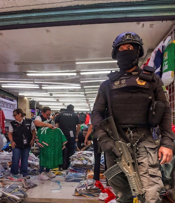 Oficial de seguridad en equipo táctico con rifle en un mercado de Tepito, mientras personal del IMPI inspecciona pilas de ropa apócrifa, incluyendo playeras