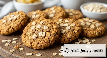 Primer plano de galletas de avena con avena esparcida sobre una tabla de madera, con cuencos borrosos de ingredientes en el fondo.
