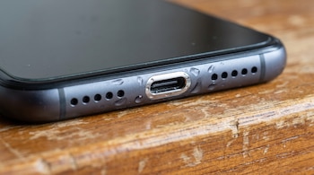 Primer plano de un teléfono móvil gris con gotas de agua en su parte inferior, incluyendo el puerto USB y los orificios de los altavoces, sobre madera.