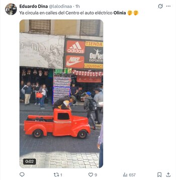 Una persona se inclina sobre una camioneta de juguete roja en una calle adoquinada, con tiendas como Adidas y Nike y transeúntes al fondo