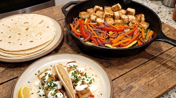 Un plato con dos tacos rellenos de pescado y verduras, un plato con tortillas y una sartén de hierro sobre una mesa de madera.