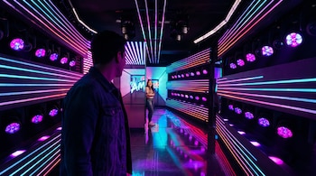 Hombre de espaldas mira a mujer sonriente al final de un pasillo. El espacio está iluminado con luces LED azules, moradas y rosas que se reflejan en el suelo.