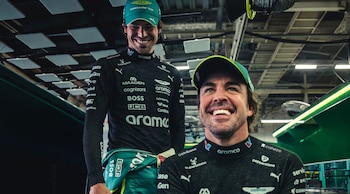 Fernando Alonso sonríe en primer plano mientras Lance Stroll permanece de pie detrás de él, ambos con vestimenta verde y negra de Aston Martin