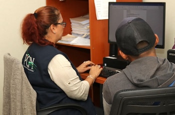 Un hombre con gafas y camisa de rayas trabaja en un escritorio; se ve una mano en el teclado y otra sosteniendo un formulario de citaciones con logo PGN