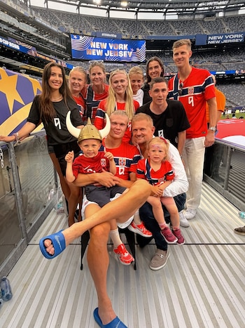 Diez personas, incluidos dos niños y Erling Haaland, posan en un estadio de fútbol. Visten camisetas de la selección de Noruega. Un niño lleva un casco vikingo. Pantalla con 'New York New Jersey'