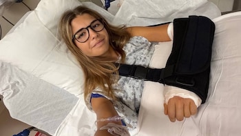 Una joven con gafas y bata médica reposa en una cama de hospital con un cabestrillo negro en el brazo izquierdo