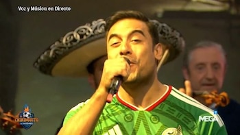 Un hombre canta en un micrófono. Lleva una camiseta verde con detalles blancos y rojos, y un escudo. Detrás, una persona con sombrero mariachi y otra con un instrumento