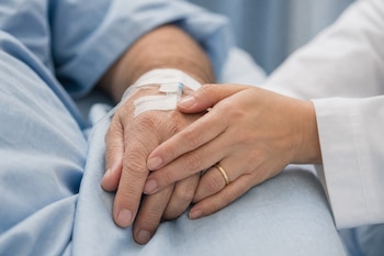 Plano detalle de una mano con vía intravenosa que es sostenida por otra mano con anillo de boda y manga blanca sobre una cama.