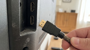 Una mano sostiene un cable HDMI trenzado con conector dorado, a punto de conectarlo a uno de los dos puertos HDMI en la parte trasera de un televisor oscuro.