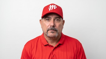 Primer plano de un hombre con bigote, gorra roja con una letra M bordada en blanco y camisa polo roja sobre un fondo blanco.