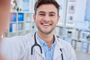 Hombre joven con pelo oscuro y barba corta, vestido con bata blanca y estetoscopio negro, sonriendo directamente a la cámara en un entorno de oficina médica
