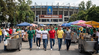 Una multitud de personas con camisetas de fútbol camina hacia el Estadio Azteca. En primer plano, vendedores ambulantes ofrecen productos en carritos metálicos.