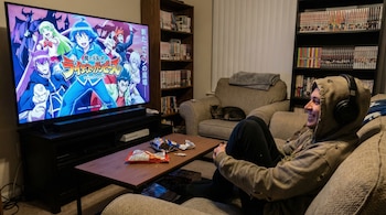 Un joven con sudadera y auriculares en un sofá mira una televisión que muestra anime. Hay una mesa con snacks y estanterías con manga. Un gato duerme.