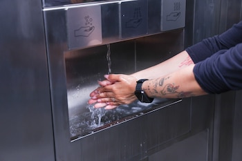 Primer plano de una persona lavándose las manos bajo un grifo de un lavabo moderno de acero inoxidable. Se aprecian manos, brazos tatuados y un reloj