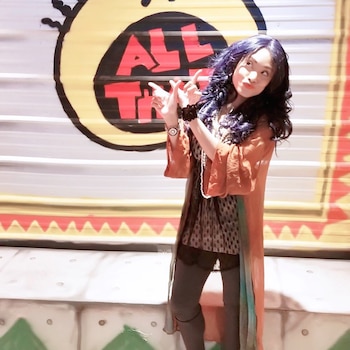 Mujer con pelo largo oscuro, cárdigan naranja, blusa estampada y reloj de pulsera. Fondo con logo circular rojo, negro, amarillo que dice "ALL THAT"