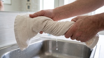 Dos manos retuercen una toalla clara sobre un fregadero metálico mientras caen gotas de agua.