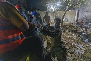 Varias personas con mascarillas, cascos y gorros, transportan escombros con cubos en un terreno con restos de construcción, bajo luz artificial nocturna