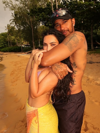 Un hombre y una mujer se abrazan en la playa. El hombre tiene tatuajes, gorra negra y gafas. La mujer lleva un bikini y falda con lentejuelas. Hay palmeras y arena