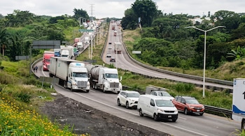 Una carretera con filas de camiones de carga, furgonetas y automóviles en movimiento entre cerros verdes.