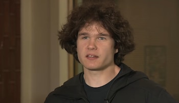 Un joven con cabello castaño rizado y brackets en los dientes viste una chaqueta negra y camiseta oscura mientras mira ligeramente a la izquierda
