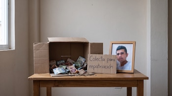 Caja de cartón con billetes y monedas sobre una mesa de madera, junto a un cartel que dice 'Colecta para repatriación' y un retrato enmarcado.