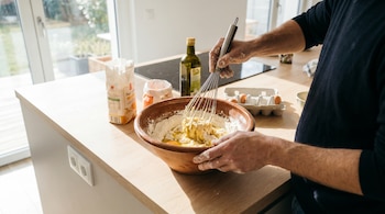 Unas manos mezclan harina, aceite y huevos en un bowl grande con un batidor de globo sobre un mesón de cocina de madera. Al fondo, harina, aceite y huevos.