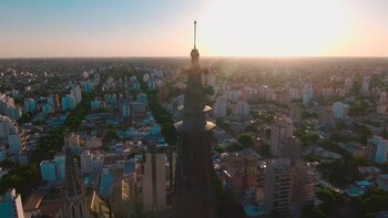 Primer plano de la aguja de la Catedral de La Plata, con un paisaje urbano extenso de edificios y árboles en el fondo bajo un cielo anaranjado al atardecer