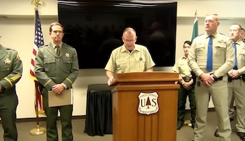 Un hombre habla en un atril con el logo del Servicio Forestal, flanqueado por seis personas en uniforme, una bandera de Estados Unidos y una pantalla oscura