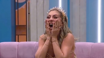 Una mujer rubia con tiara brillante, vestido dorado y manos en las mejillas, sentada en un sofá rosa.