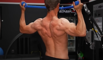 Jeff Cavaliere, de espaldas y sin camisa, realiza un ejercicio con una cuerda azul conectada a una máquina de gimnasio, mostrando su musculosa espalda