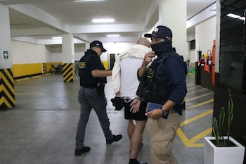 Dos agentes policiales custodian a un hombre esposado que se cubre el rostro en un estacionamiento subterráneo