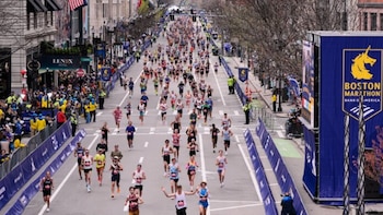 Cientos de corredores avanzan por una calle ancha durante el Maratón de Boston, con edificios y espectadores a los lados, y una gran pancarta azul de la carrera