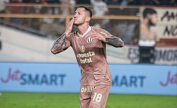 Hombre con camiseta de fútbol marrón número 18, tatuajes en los brazos, sopla un beso. Fondo con tribunas y banner publicitario borrosos