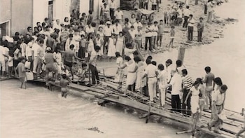 Un grupo de personas camina sobre un puente improvisado de madera, que se extiende sobre un río crecido. Varias personas observan desde la orilla