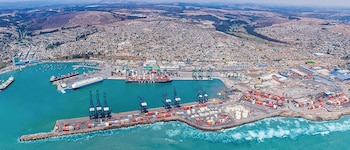 Vista aérea de un gran puerto con varios barcos de carga, grúas y pilas de contenedores junto a una ciudad costera densamente poblada con colinas de fondo