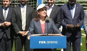 Mujer hablando en un podio azul con 'FIFA arena', vistiendo un blazer y una visera blanca, con varios hombres en traje detrás de ella en un día soleado