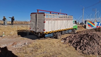 Dos agentes de policía con equipo táctico junto a un barril rotulado "POLICIA" en el hombro de una carretera, con un camión blanco y verde y un cielo azul despejado