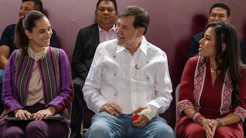 Tres personas sentadas conversan y sonríen en un acto público, incluyendo a un hombre con camisa blanca y vendaje en la mano izquierda