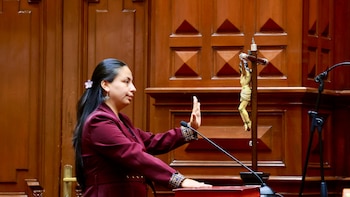 Mujer en traje color borgoña con mano derecha levantada y mano izquierda sobre un libro rojo. Al fondo, pared de madera y un crucifijo con un micrófono