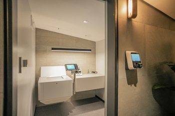 Sala moderna con paredes de azulejos, escritorio flotante, terminal con pantalla, mueble blanco, alfombra gris, luz lineal de techo. Puerta blanca y dispositivo en pared externa